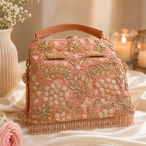 May include: A peach-colored handbag featuring intricate gold thread embroidery in floral and leaf patterns, accented with small white beads. The bag has a top handle, a drawstring closure, and a bottom fringe made of delicate hanging beads.