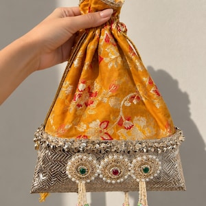 May include: A drawstring pouch featuring a mustard yellow fabric upper with a floral pattern in red and silver. The base is a metallic silver woven material adorned with three circular embroidered medallions, each featuring a central gemstone.