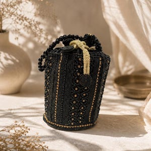 May include: A cylindrical bucket bag covered in intricate black beadwork and gold metallic accents. The bag features a drawstring closure with a gold braided cord and a handle made of large, round black beads. The surface displays vertical patterns.