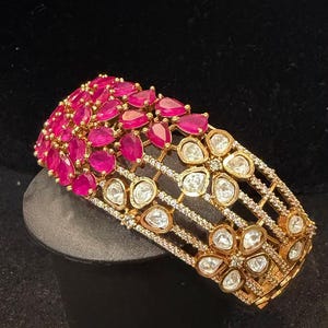 May include: A wide gold-tone cuff bracelet featuring a dense cluster of pear-shaped fuchsia pink stones on one side and a series of openwork floral motifs set with clear, irregular-shaped stones and small pavé accents across the remaining band.
