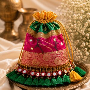 May include: A drawstring pouch featuring a vibrant magenta body with gold floral patterns, a green upper section, and a yellow drawstring top. The base is adorned with a row of small circular mirrors and a fringe of green and yellow tassels.