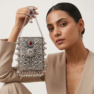 May include: A rectangular silver-toned metal pouch featuring intricate floral engravings, a central oval red stone, and a chain handle with a matching red stone ring attachment. The pouch is adorned with small dangling silver bells along the sides and base.