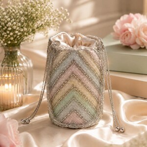 May include: A cylindrical drawstring pouch bag featuring a chevron pattern of pastel-colored beads in shades of pink, yellow, green, and blue, accented with silver metallic thread and silver-toned metal beads, finished with two silver chain drawstrings.