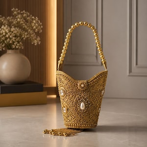May include: A gold-toned bucket-style handbag featuring an intricate filigree lace pattern, embellished with central teardrop-shaped faux pearls and circular accents. The bag includes a beaded handle and a dangling tassel of gold chains and spheres.