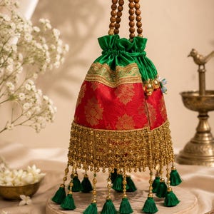 May include: A drawstring pouch bag featuring a red fabric body with a gold floral pattern, a green satin top section, and a handle made of brown wooden beads. The base is adorned with gold metallic fringe and hanging green thread tassels.