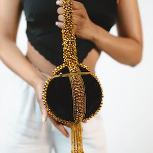May include: A black velvet handbag with gold beaded accents and a gold chain handle. The round bag features a vertical gold band and a gold tassel. The bag is held by a person.