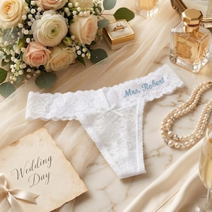 May include: White lace thong underwear with the text "Mrs. Robert" embroidered in blue. The underwear is displayed with a bouquet of roses, a wedding ring, perfume, and a "Wedding Day" card.