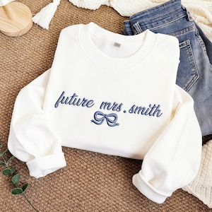 Könnte beinhalten: Ein weißes Sweatshirt mit dem blauen Schriftzug "future mrs. smith" und einer blauen Schleife. Das langärmlige Oberteil ist aus einem weichen Material gefertigt und eignet sich für den täglichen Gebrauch.