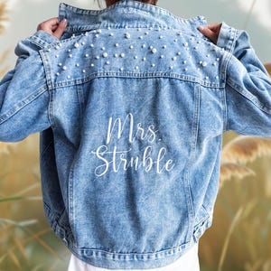 May include: Light blue denim jacket with pearl embellishments on the upper back. The jacket features the embroidered text "Mrs. Struble" in white script. The date "06.24.2023" is embroidered on the collar.