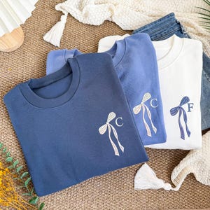 Op de afbeelding: Drie crewneck sweatshirts in blauw- en wittinten. Elk sweatshirt heeft een geborduurd strikdesign met een enkele initiaal. De sweatshirts zijn opgevouwen en op elkaar gestapeld, waarbij de verschillende kleuren en ontwerpen worden getoond.