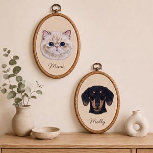 May include: Two oval wooden embroidery hoops containing fabric portraits of pets. The top hoop features a cream-coloured cat with blue eyes and the name Mimi embroidered below. The bottom hoop shows a black and tan dachshund with the name Molly.