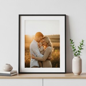 Może przedstawiać: Oprawiony portret rodzinny przedstawiający parę obejmującą dziecko na polu o zachodzie słońca. Czarna rama otacza fotografię, która ma białe passe-partout. Rodzina ubrana jest w ubrania w neutralnych odcieniach.