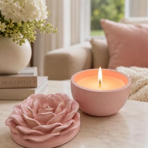 May include: A light pink candle in a round container with a matching lid shaped like a blooming rose. The candle is lit, displaying a bright flame and warm wax. The rose-shaped lid rests beside the candle, showing detailed petal layers.