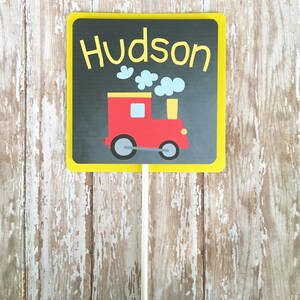 May include: A yellow sign with black chalkboard background and a red toy train with black wheels. The sign says "Hudson" in yellow letters.