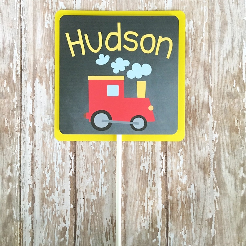 May include: A yellow sign with black chalkboard background and a red toy train with black wheels. The sign says "Hudson" in yellow letters.