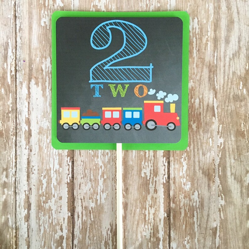 May include: A green chalkboard sign with a blue number two and the word "TWO" written in colorful letters. A toy train with red, yellow, blue, and green cars is pictured below the number.