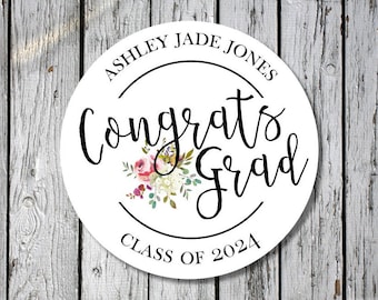 Puede incluir: Señal redonda blanca con texto negro que dice "Ashley Jade Jones Felicidades Graduado Clase de 2024". La señal tiene un diseño floral con flores rosas, blancas y verdes.