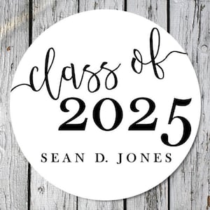 May include: A white circular sticker with black text. The words "class of" are in a cursive font above the bold numbers "2025". Below the numbers is the name "SEAN D. JONES" in a simple, sans-serif font. The background is a weathered wood.