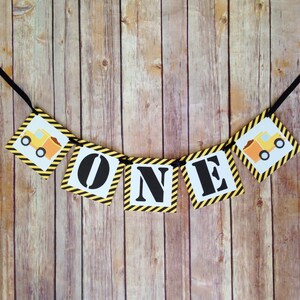 May include: A yellow and black construction themed banner with the word "ONE" in black letters. The banner has small images of yellow dump trucks with black wheels.