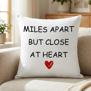 May include: White square pillow with the text "MILES APART BUT CLOSE AT HEART" in black, with a red heart below. The pillow is on a beige sofa, with plants and a window in the background.