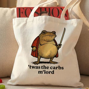 以下が含まれることがあります： 赤いマントを着て剣を持ったカエルの漫画が描かれた白いキャンバスのトートバッグ。カエルの下には「'twas the carbs m'lord」というテキストが印刷されています。バッグには2つのハンドルがあり、雑誌が入っています。