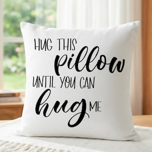 Puede incluir: Cojín cuadrado blanco con el texto "HUG THIS pillow UNTIL YOU CAN hug ME" en escritura negra. El cojín está sobre una mesa de madera con un camino de mesa blanco.