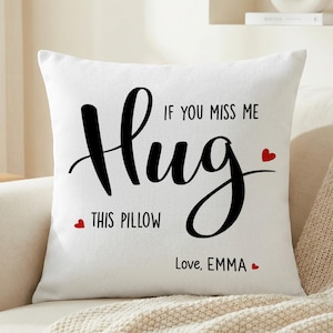 Puede incluir: Cojín cuadrado blanco con la palabra "Hug" en una fuente cursiva grande y negra. El texto "If you miss me, this pillow" está arriba y abajo de la palabra "Hug". Pequeños corazones rojos acentúan el texto. Las palabras "Love, EMMA" están en la parte inferior.