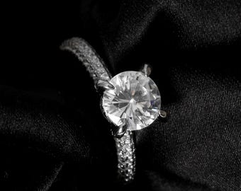 May include: A silver ring with a large, round, clear gemstone as the centerpiece. The band is encrusted with smaller, sparkling stones. The ring is set against a black, silky background, with white flowers in the upper left corner.