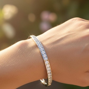 Pode incluir: Uma pulseira de prata com numerosas pedras preciosas redondas e transparentes. A pulseira é usada no pulso de uma pessoa, com as pedras preciosas captando a luz. O fundo está desfocado, com toques de verde e amarelo.
