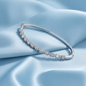 May include: A silver bracelet featuring a line of clear, sparkling gemstones. The bracelet is displayed on a light blue satin background, which enhances the sparkle of the stones. The clasp is visible. The bracelet is approximately 17 cm in length.