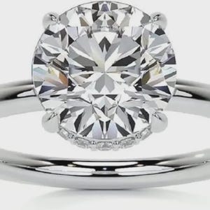 May include: A silver engagement ring with a large, round, clear diamond as the centerpiece. The diamond is set with four prongs and surrounded by smaller accent stones. The band is smooth and polished.
