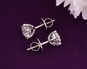 May include: A pair of silver stud earrings featuring clear, round-cut gemstones. The earrings are set in a prong setting, and the posts have a butterfly backing. The earrings are presented on a deep purple background.