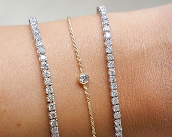 May include: Three delicate bracelets. Two are silver-toned, each lined with small, sparkling stones. The third is gold-toned, featuring a single, round, clear stone. The bracelets are displayed on a wrist.