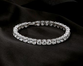 May include: A silver tennis bracelet with numerous round, clear gemstones. The bracelet is set on a black, textured background, creating a contrast that highlights the sparkle of the stones. The clasp is visible.