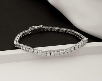 May include: A silver bracelet with a row of square-cut, clear gemstones. The bracelet is set against a white and black background, with a white fabric draped in the background. The clasp is visible.