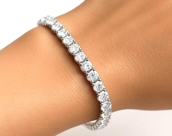 May include: A tennis bracelet with multiple round, clear gemstones set in a silver-colored metal. The bracelet is worn on a wrist, showcasing the sparkle of the stones. The bracelet is a classic piece of jewelry.