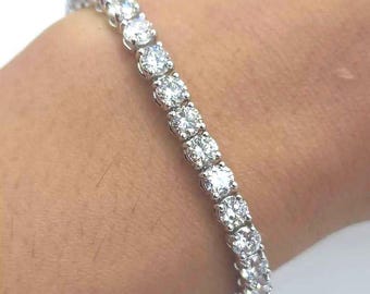 May include: A tennis bracelet featuring a series of round, clear gemstones set in a silver or white metal band. The gemstones are evenly spaced and reflect light, creating a sparkling effect. The bracelet is worn on a wrist.