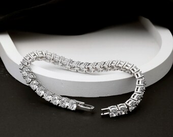 May include: A silver tennis bracelet with a row of round, clear gemstones. The bracelet is unclasped, revealing the clasp mechanism. The jewelry is displayed on a white, curved tray against a black background.