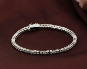 May include: A silver tennis bracelet with a row of small, clear gemstones. The bracelet is set against a dark red-brown background, with a secure clasp. The gemstones are evenly spaced and reflect light.