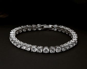 May include: A silver tennis bracelet with a row of round, clear gemstones. The bracelet is set against a black background, highlighting the sparkle of the stones. The clasp is visible.
