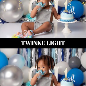 May include: A child in a grey romper sits in front of a backdrop with balloons and a cake. The child has blue frosting on their face and hands. A small cake with a number 1 on top sits on a white cake stand. The word "TWINKE LIGHT" is in the image.
