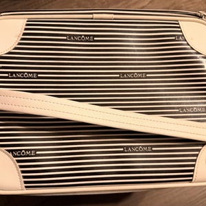 Puede incluir: Un neceser rectangular con un estampado de rayas blancas y negras. La bolsa tiene detalles de cuero blanco en las esquinas y un asa blanca. La palabra "LANCÔME" está impresa en la bolsa. Un pequeño logotipo circular blanco está en la esquina inferior derecha.