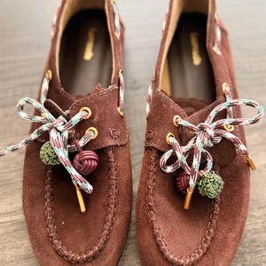 May include: A pair of brown suede loafers with decorative laces. The laces are multi-coloured with green and burgundy pom-poms. The shoes have gold eyelets and a stitched design around the toe.