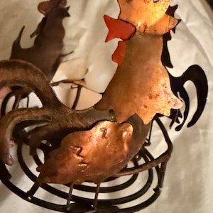 May include: A metal decorative holder featuring three rooster silhouettes arranged in a circle. The roosters are crafted from copper-toned metal with dark brown accents, and the base is constructed from a circular wire coil design.