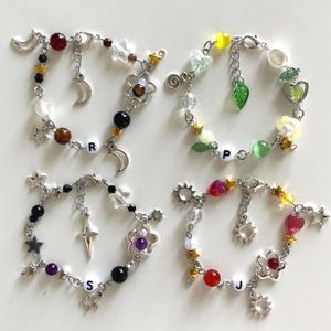 May include: Four handmade charm bracelets with silver chains, featuring various beads, charms, and letter beads. Each bracelet has a different color scheme, including red, black, purple, and yellow beads, with star, moon, and flower charms.