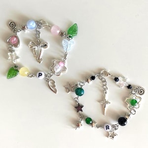 Può includere: Due braccialetti di perline con catene color argento. Uno presenta perline rosa, blu e verdi, un ciondolo a forma di croce e la lettera "P". L'altro ha perline verdi e nere, ciondoli a stella, una croce e la lettera "R".