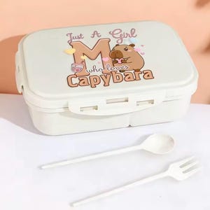 May include: A light beige lunchbox with a cartoon capybara design and the text "Just A Girl who loves Capybara". Includes a white spoon and fork. The lunchbox has a secure latch.