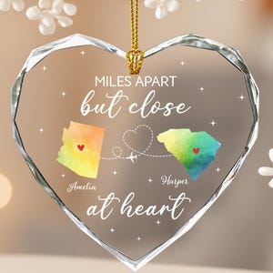 May include: A heart-shaped glass ornament with a gold chain. The ornament features two colorful state maps, a heart, and the text "MILES APART but close at heart." The names "Amelia" and "Harper" are also included.