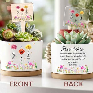 May include: White ceramic planter with a wooden base, featuring a succulent arrangement and floral designs. The front of the planter has the names Taylor, Tina, and Sophia. The back has the word "Friendship" and a quote. A wooden sign with floral designs is in the arrangement.