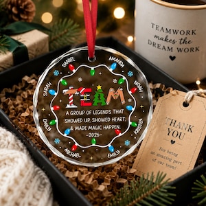 May include: A round, clear glass Christmas ornament with a red ribbon hanger. The design features a string of colorful holiday lights, the word TEAM, and the text: A group of legends that showed up, showed heart, & made magic happen. -2026-